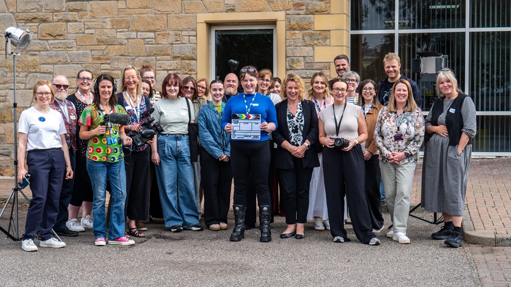 A large group of people standing in front of a building. The person in the centre holds a clapper board.