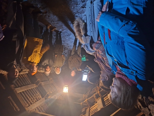 A group in warm outdoor clothing gathered inside a dimly lit, rustic building with exposed stone walls, a thatched roof, and a central hanging light.