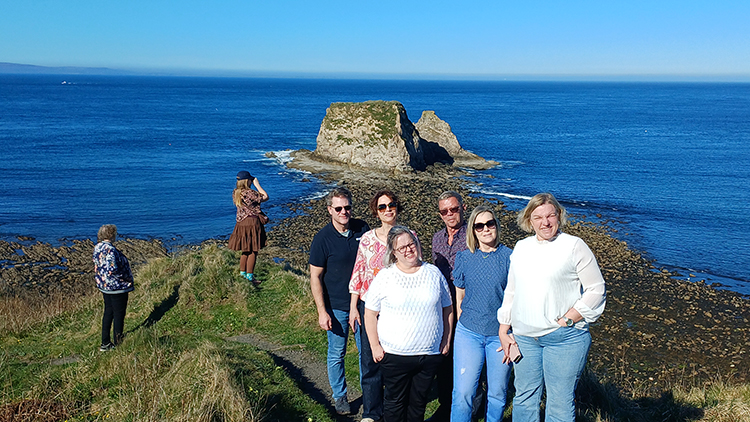 A group of people standing posing for a photo against a clifftop on a sunny day with the sea and sea stacks visible in the background A group of people standing posing for a photo against a clifftop on a sunny day with the sea and sea stacks visible in the background