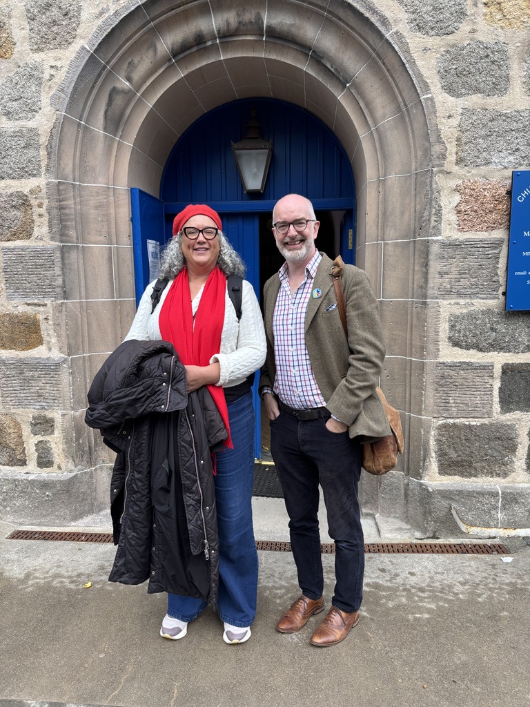 The photo shows two people standing in front of a stone building with a blue arched door. The person on the left is wearing a red hat, red scarf, white top, and blue jeans, and is holding a black coat. The person on the right is wearing a brown jacket, checked shirt, and dark trousers with brown shoes. Both are smiling.