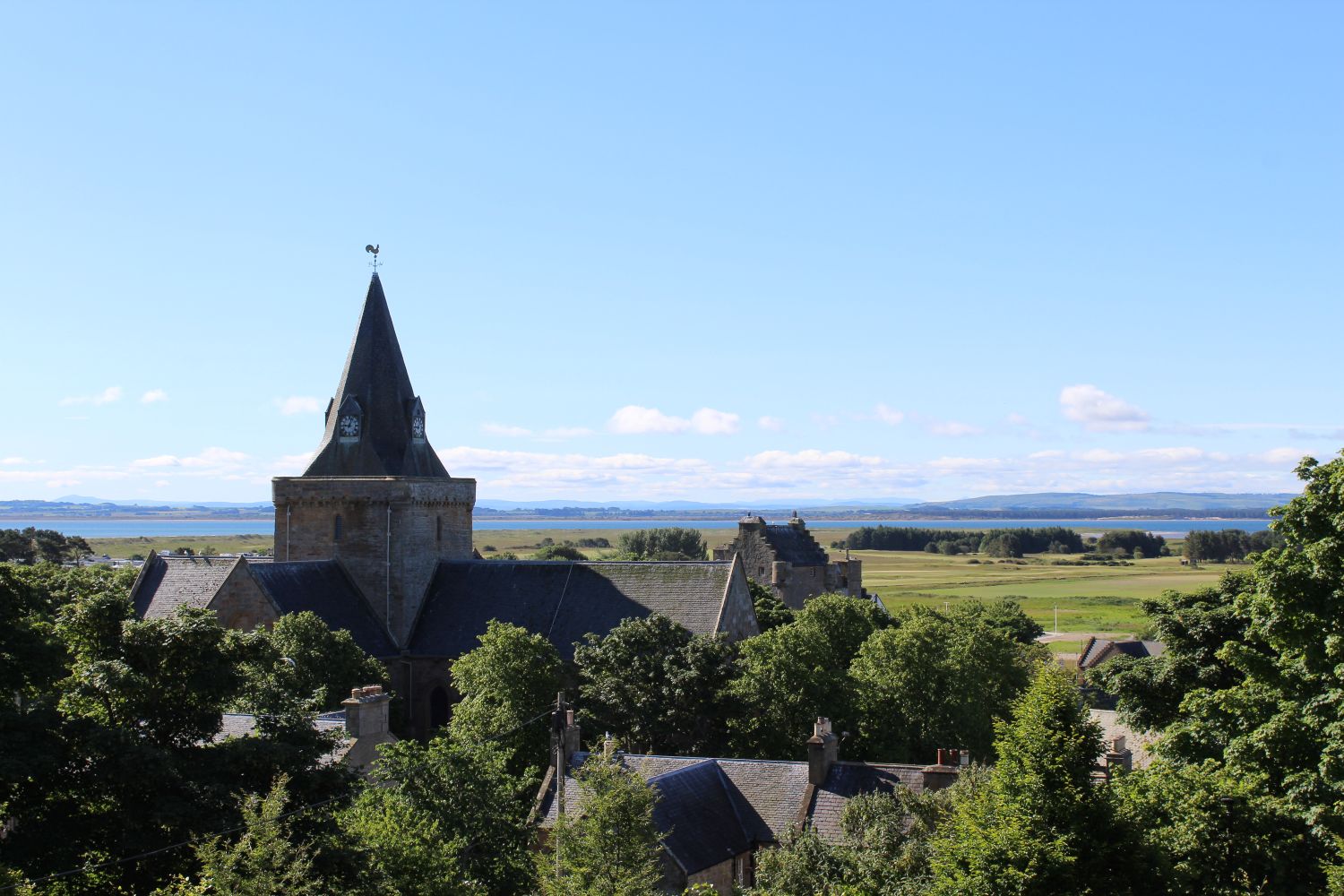 Townscape of a cathedral spire and roofs of houses, surrounded by leafy trees with green grasslands and the blue sea in the background