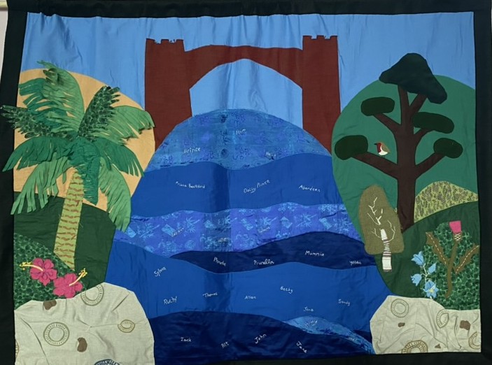 A textile piece depicting a blue river flowing beneath a large brown bridge, surrounded by green trees and plants on both sides, with a light blue sky in the background.