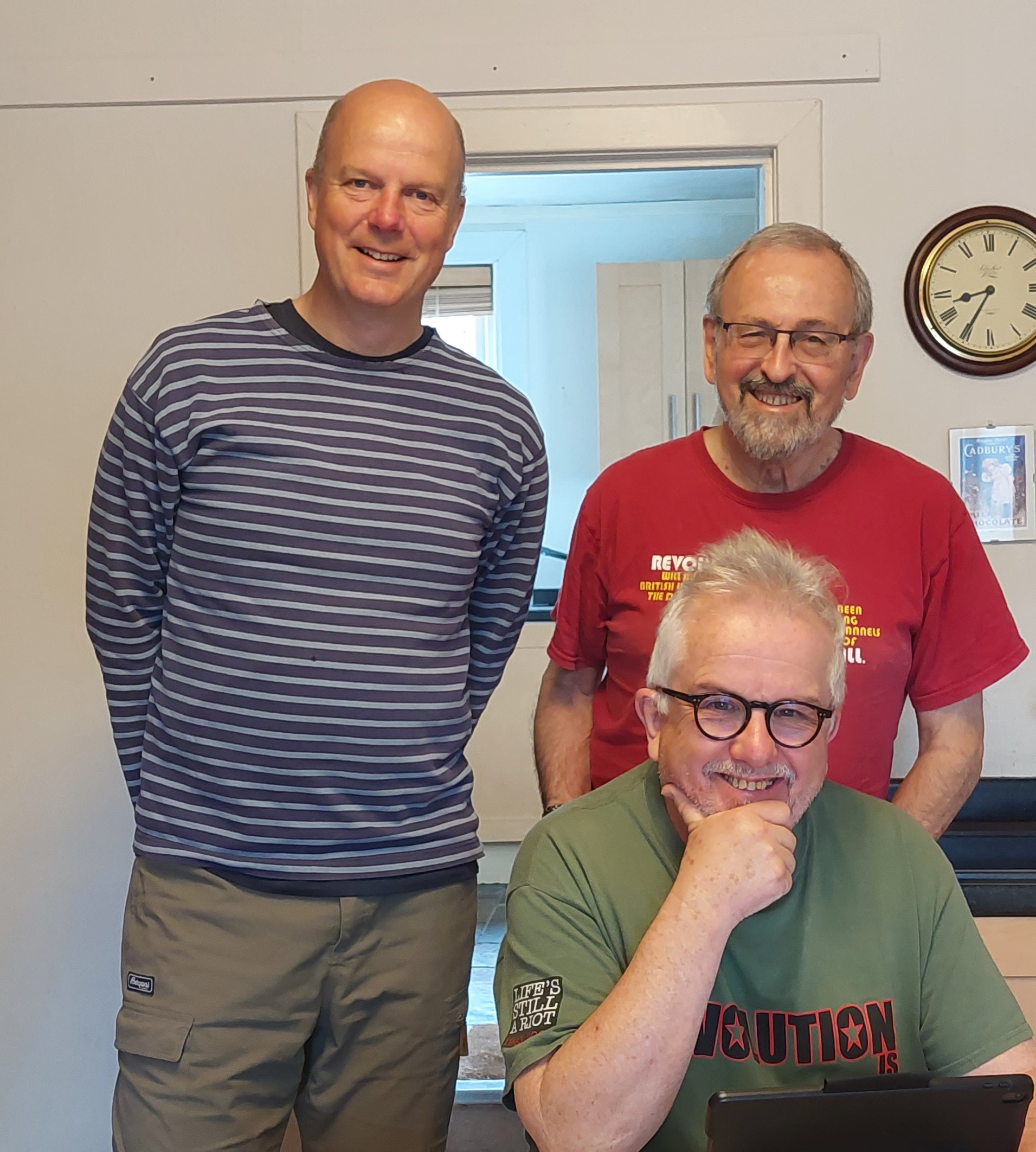 Two men standing with a third one seated in the foreground. All smiling at the camera, gathered together as a close group Two men standing with a third one seated in the foreground. All smiling at the camera, gathered together as a close group