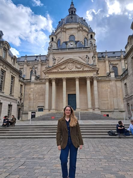 Woman standing on the steps leading towards a grand building with Corinthian columns supporting a decorated pediment, leading up towards a dome adorned with statues and a clockface. Woman standing on the steps leading towards a grand building with Corinthian columns supporting a decorated pediment, leading up towards a dome adorned with statues and a clockface.