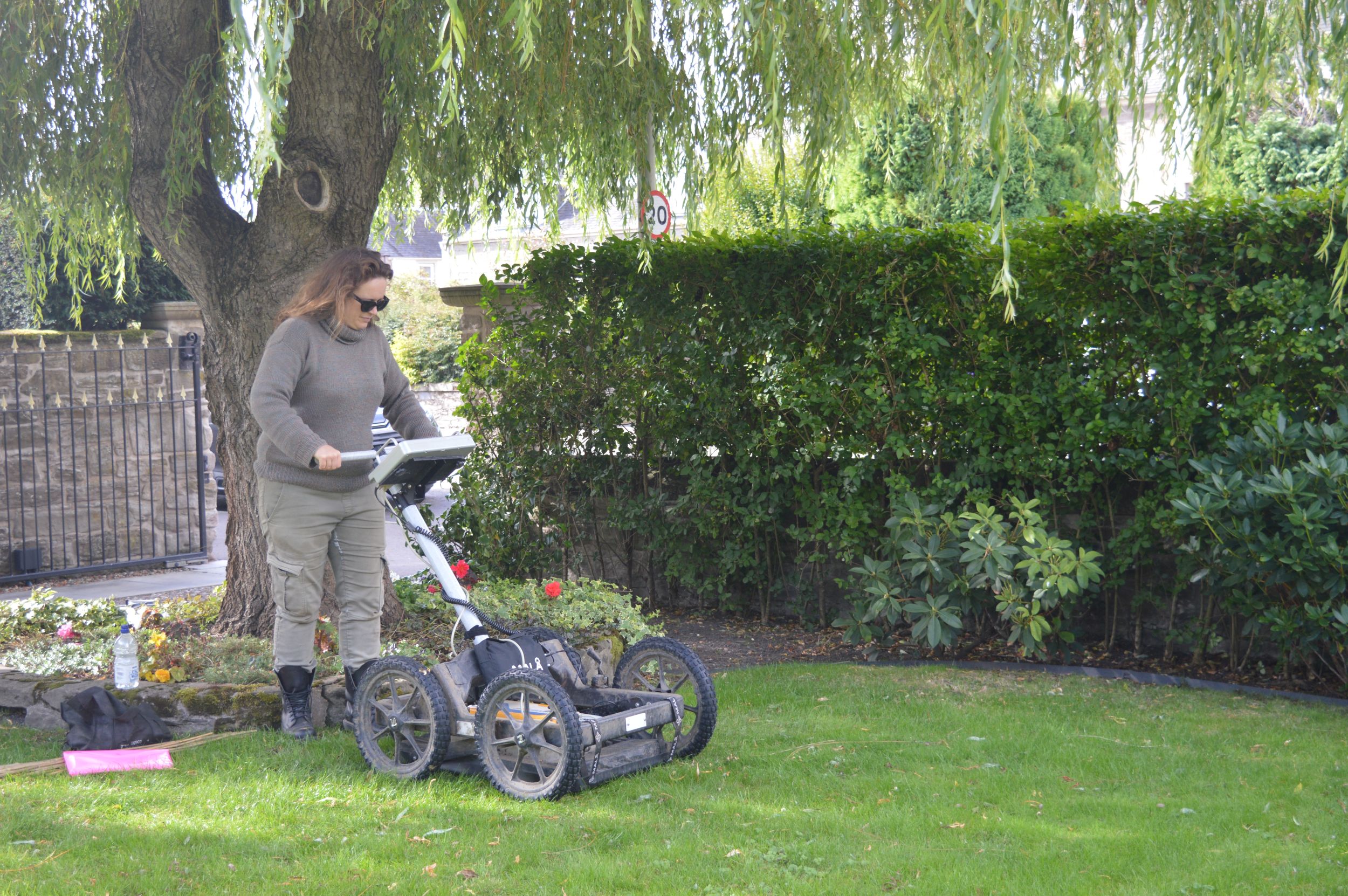 Woman operating a ground penetrating radar scanner in a grassy area with a tree and a hedge framing it