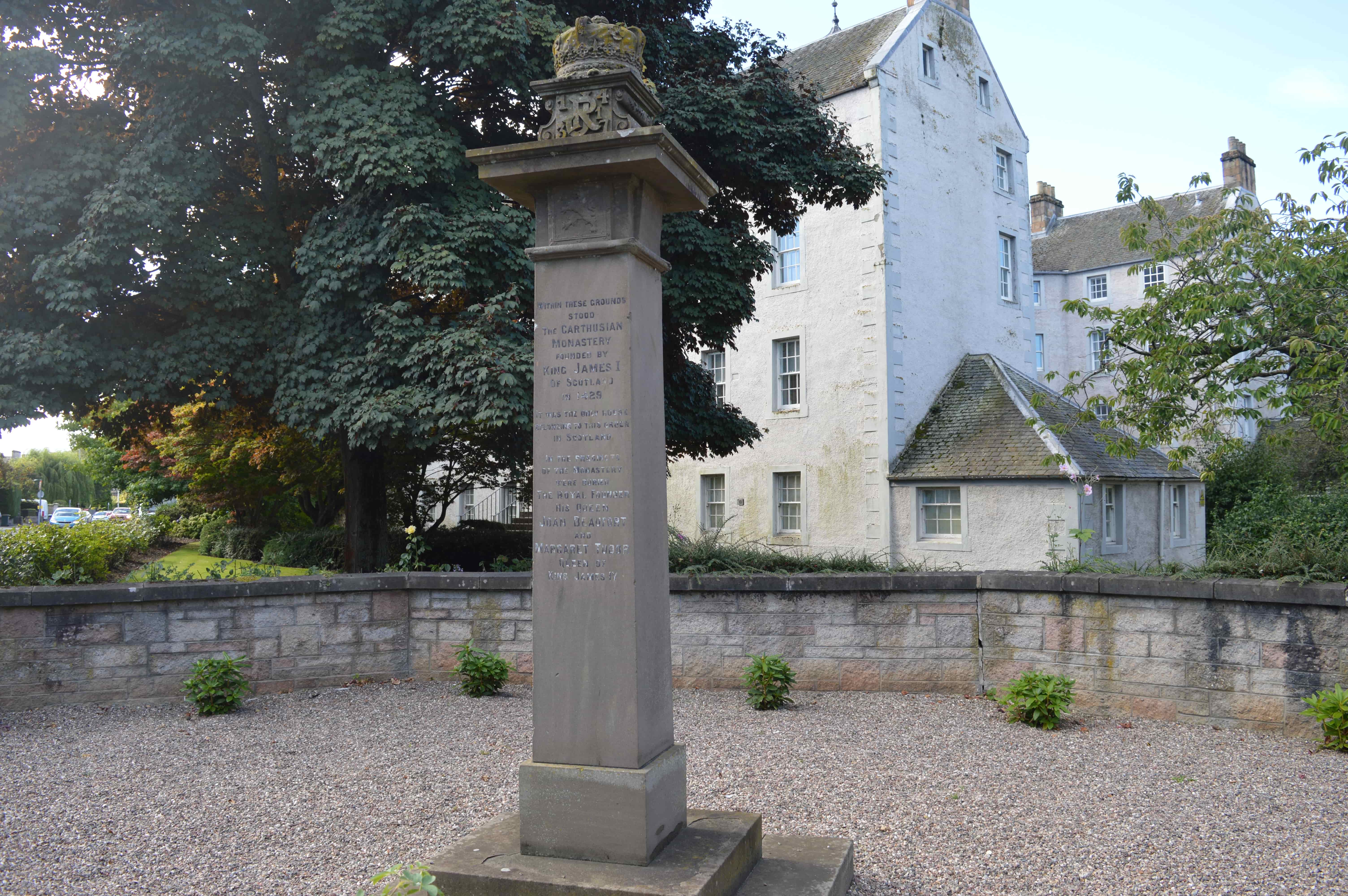 Pillar-style stone monument, with a gravelled area around it. In the background there are leafy trees and a tall stone building
