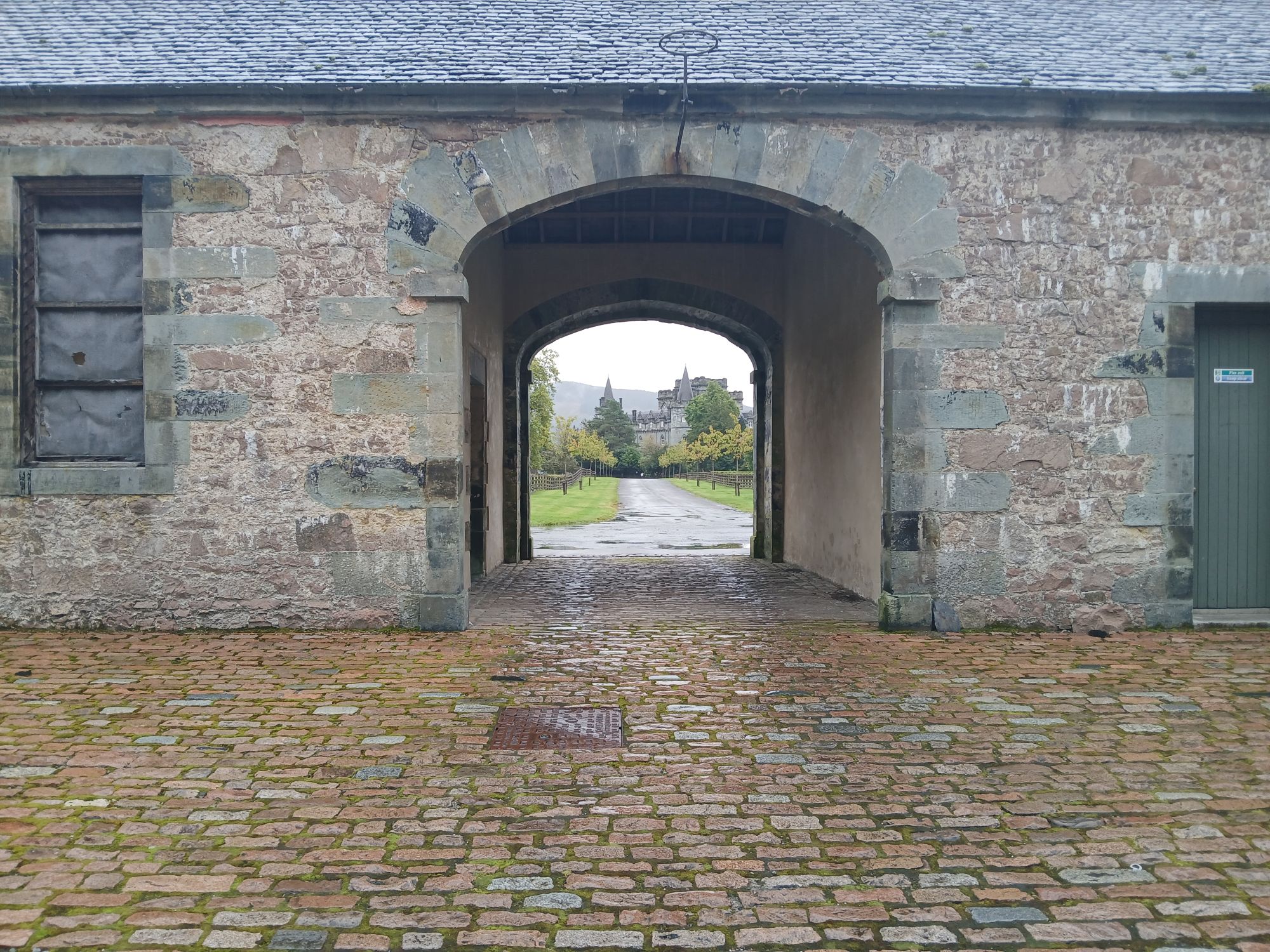 A thick stone wall with an arched gateway through it, with a view of a castle seen through the arch.