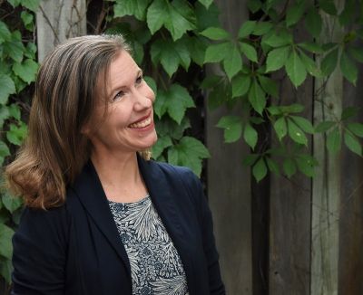 A smiling woman standing in the foreground of the image, with a fence with leafy vines behind her. A smiling woman standing in the foreground of the image, with a fence with leafy vines behind her.