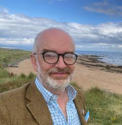 Close up of a man with a beard and glasses, wearing a light blue shirt and brown tweed jacket. Background shows a sandy beach and sea, with blue sky. 