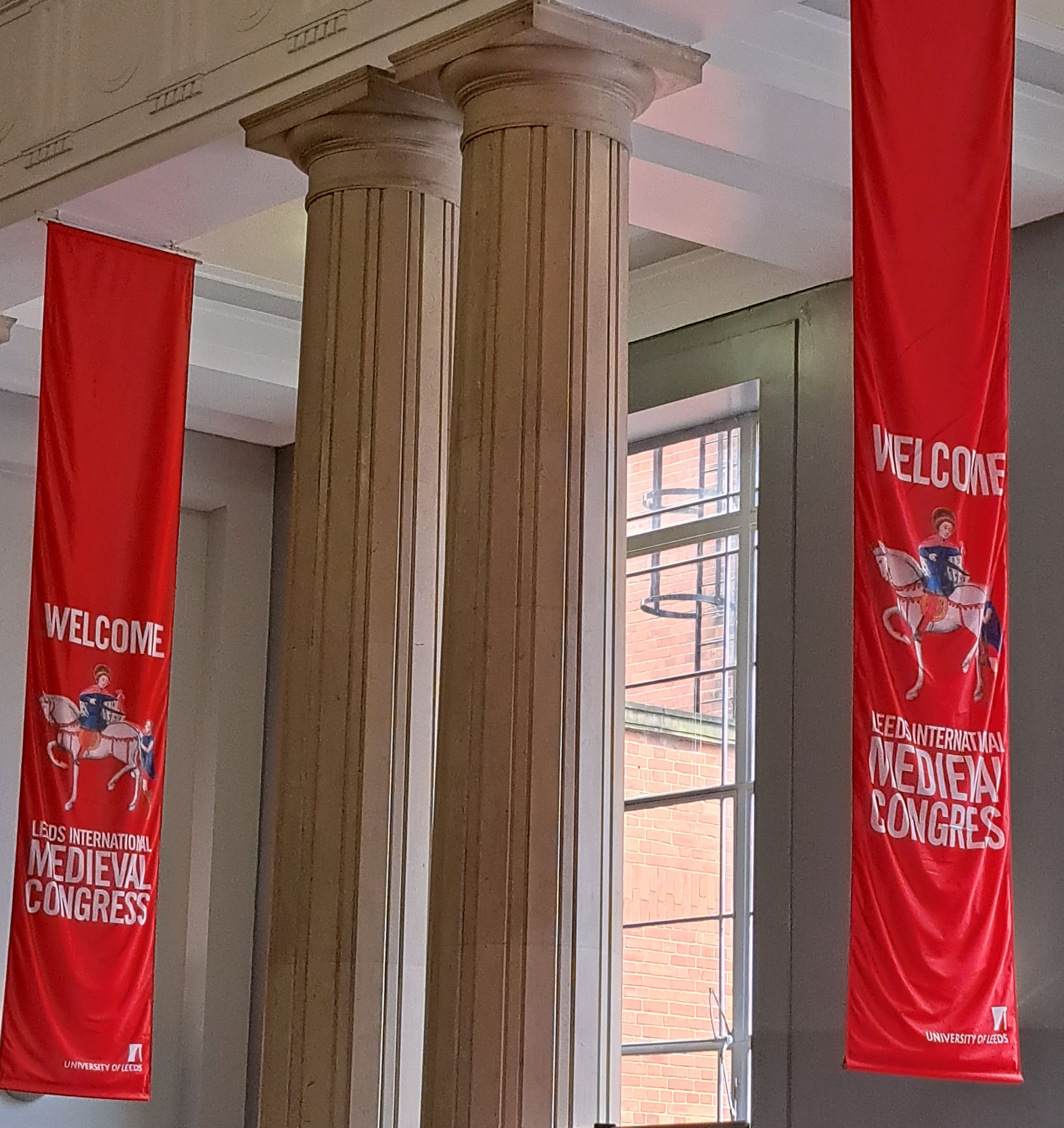 Red banners hung from the ceiling with picture of a medieval manuscript-style knight on horseback and text of 'Welcome, International Medieval Congress'. In between the two banners, there are two classical columns. Red banners hung from the ceiling with picture of a medieval manuscript-style knight on horseback and text of 'Welcome, International Medieval Congress'. In between the two banners, there are two classical columns.