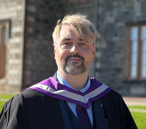 Man wearing graduation robes and a hood, with a stone building behind him. 