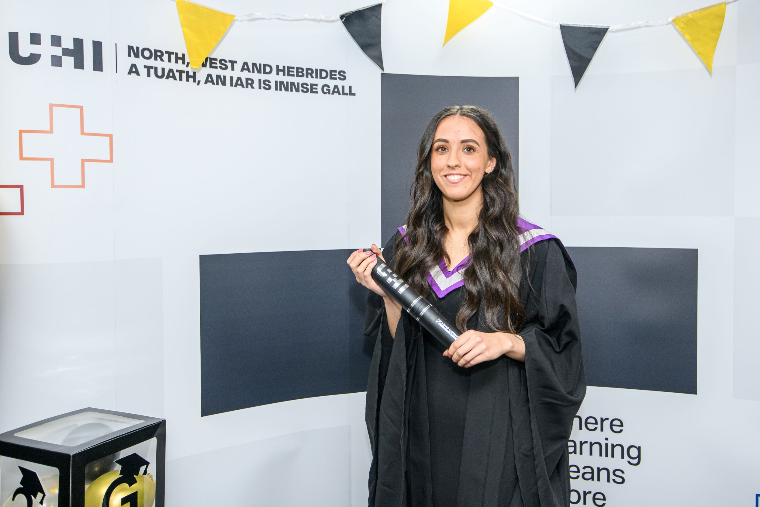 Woman with long dark hair wearing a black dress, a graduation robe and hood, holding a degree scroll. Above her, there is gold and black bunting.