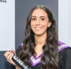 A smiling woman wearing a graduation gown and hood, holding a degree scroll